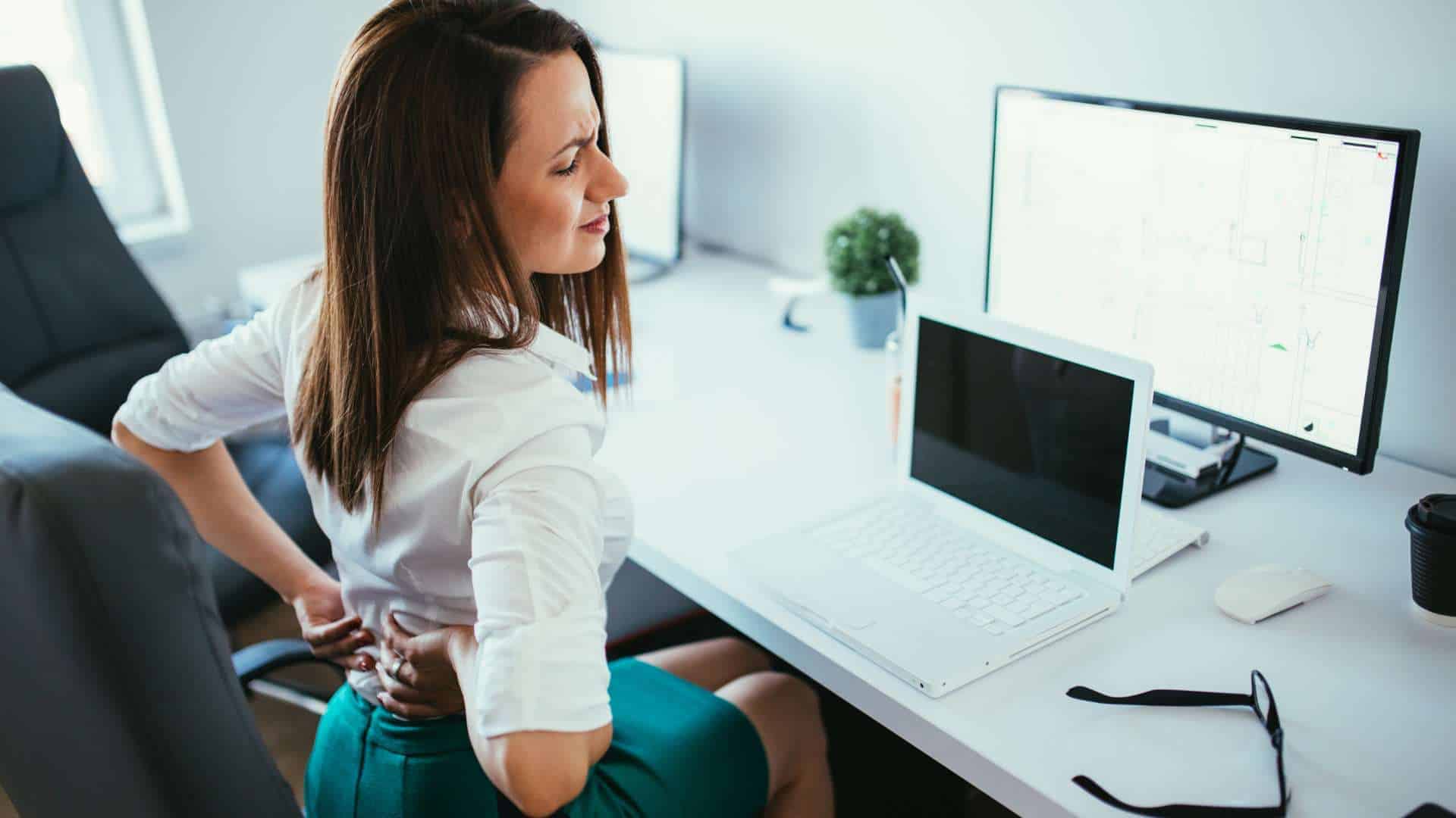 A woman sitting at a desk in an office setting grimaces and holds her lower back, suggesting discomfort. Sporting a white blouse and teal skirt, she sits near a laptop, monitor, glasses, and potted plant. She might consider pain management NYC or physical therapy NYC for relief.