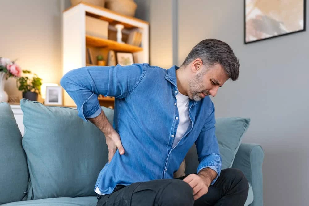 A man in a blue denim shirt and white t-shirt sits on a teal sofa at home, clutching his lower back with a pained expression. Considering physical therapy NYC might offer the relief he needs. A bookshelf with various items is in the background.