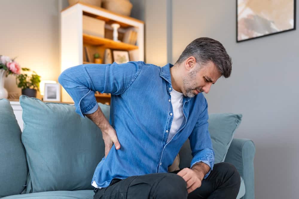A man in a blue denim shirt and white t-shirt sits on a teal sofa at home, clutching his lower back with a pained expression. Considering physical therapy NYC might offer the relief he needs. A bookshelf with various items is in the background.