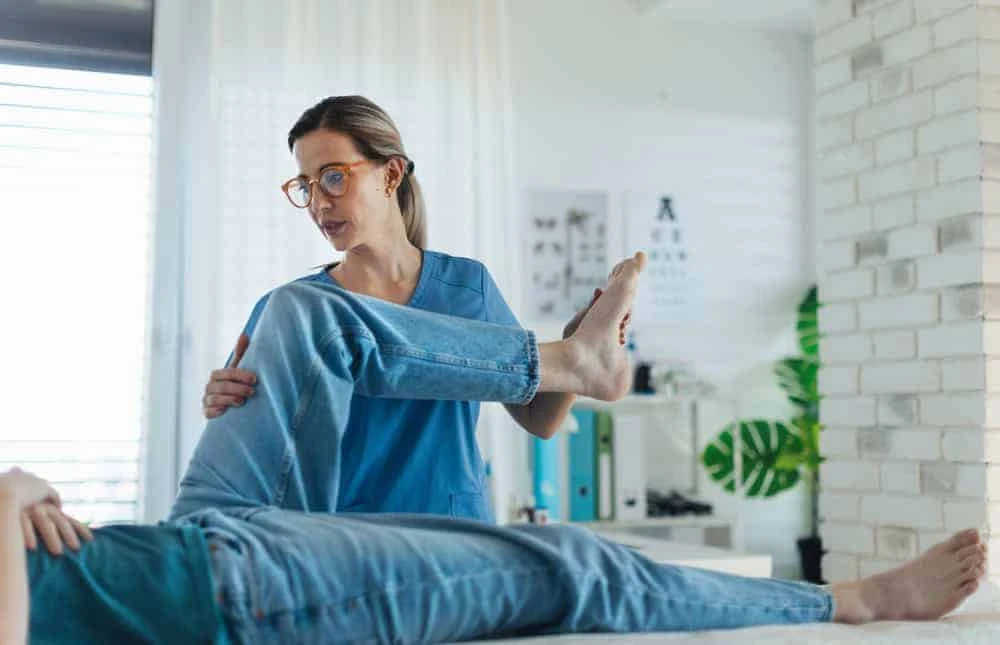 A healthcare professional in blue scrubs assists a person lying down by lifting and bending their leg at the knee. The setting, suggesting a physical therapy NYC clinic, features a plant and various charts in the background.
