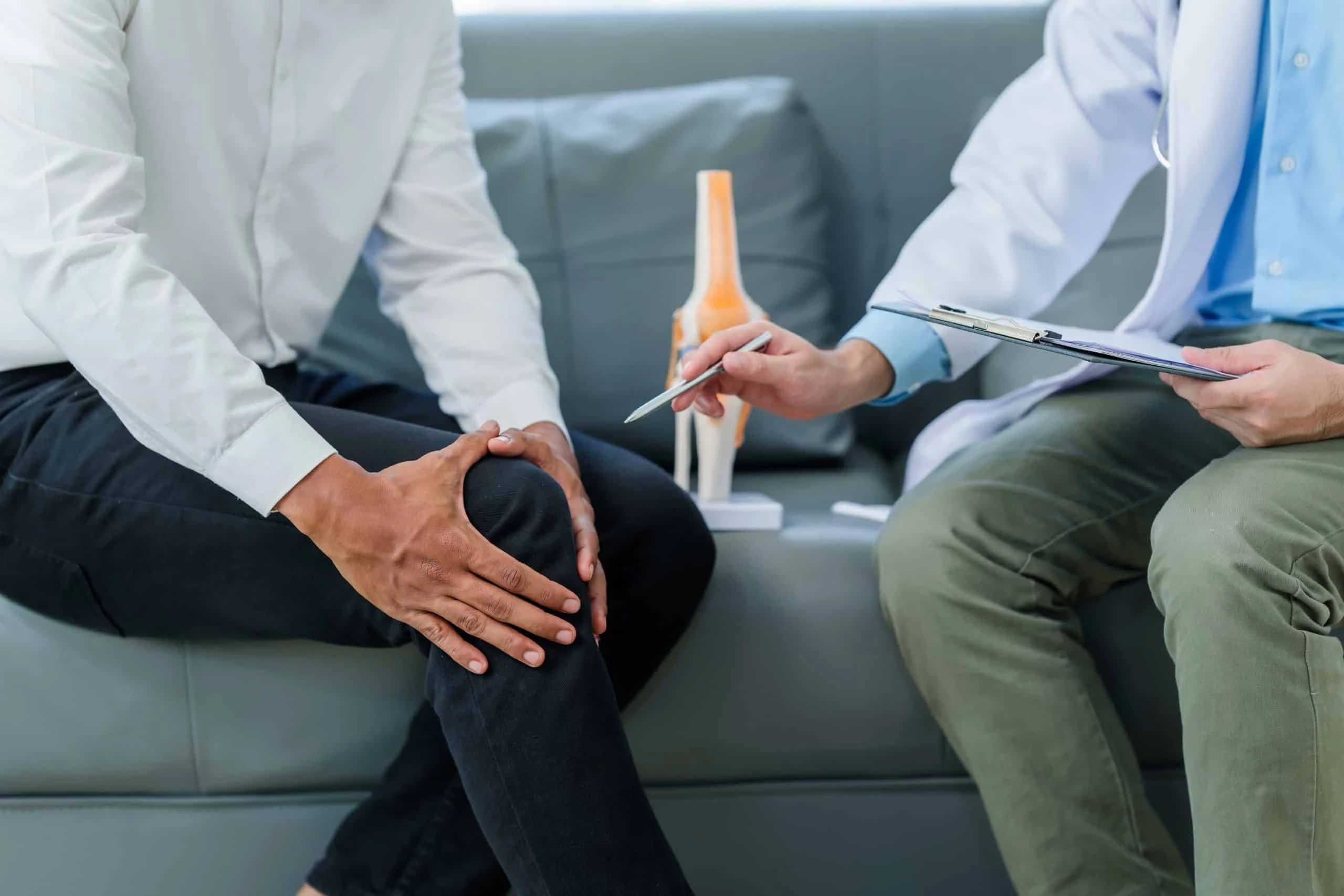 A person holds their knee while sitting on a couch, discussing pain management NYC with a doctor who is pointing at a clipboard. A model of a knee joint is visible in the background, highlighting the importance of physical therapy NYC for rehabilitation.