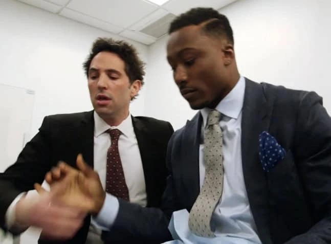 In a brightly lit NYC office, two men in suits are shaking hands. The man on the left, with dark hair in a dark suit, converses with the right-side counterpart who sports a tie and pocket square. Both appear engaged, as if discussing pain management or physical therapy strategies.