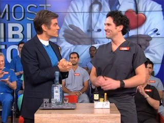 Two men stand and smile at each other on a television set with a medical theme. One man wears a suit, the other is in scrubs, discussing pain management NYC. A table with a bottle and some items separates them, while an audience clad in scrubs is visible in the background.