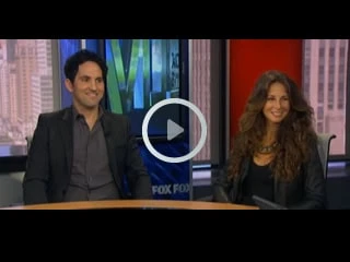 A man and a woman are sitting at a news studio desk, smiling. The backdrop features a cityscape with the words "FOX FOX," while a play button symbol floats in the center, inviting viewers to explore topics like physical therapy NYC and pain management NYC.