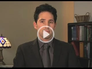 A man in a suit stands indoors, facing the camera. Behind him is a bookshelf filled with books and a decorative basket. A stained-glass lamp can be seen on the left side. The image features a play button over it, likely indicating expertise in pain management NYC through video insights.