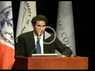 A person is standing at a podium, speaking into a microphone. Behind them are various flags and a banner, suggesting a formal event or lecture possibly discussing innovations in physical therapy NYC.