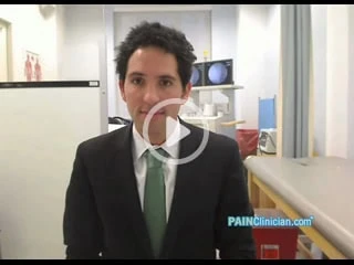 A man in a suit and green tie stands in a clinical setting, surrounded by medical equipment, emphasizing his expertise in pain management NYC. The logo "PAINClinician.com" is visible on the lower right, with a play button icon overlaid on the image.