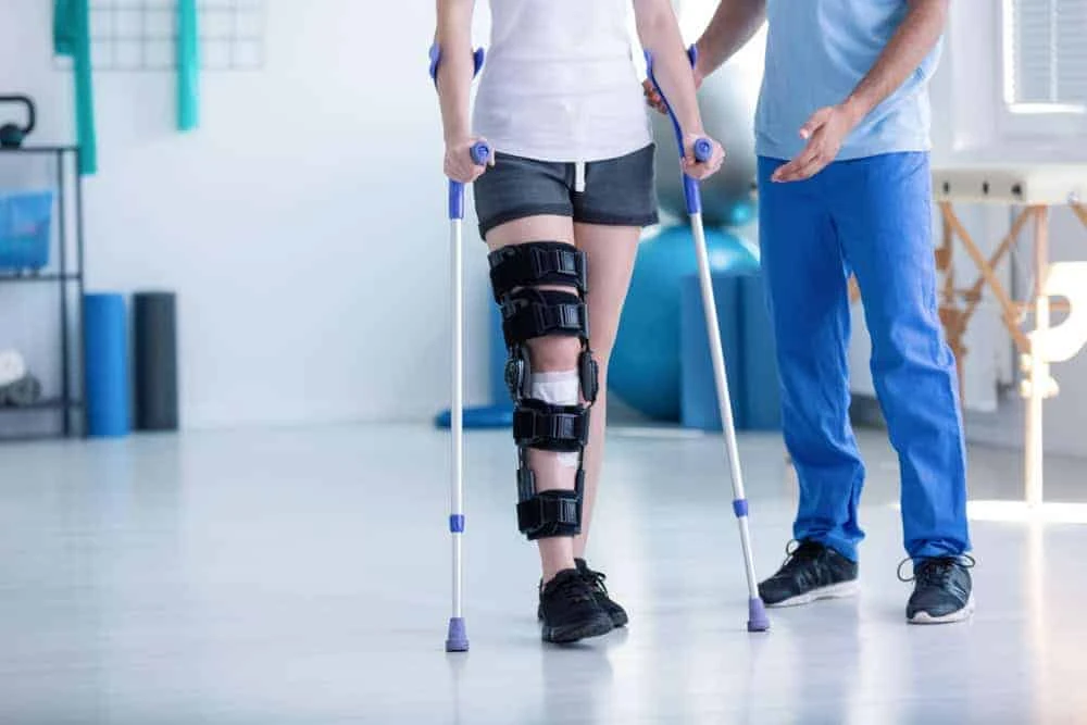 In a bright rehabilitation room, guided by a therapist in blue scrubs, a person on crutches with a leg brace takes a step forward. Physical therapy NYC techniques are at play, promising effective pain management with exercise equipment visible in the background.