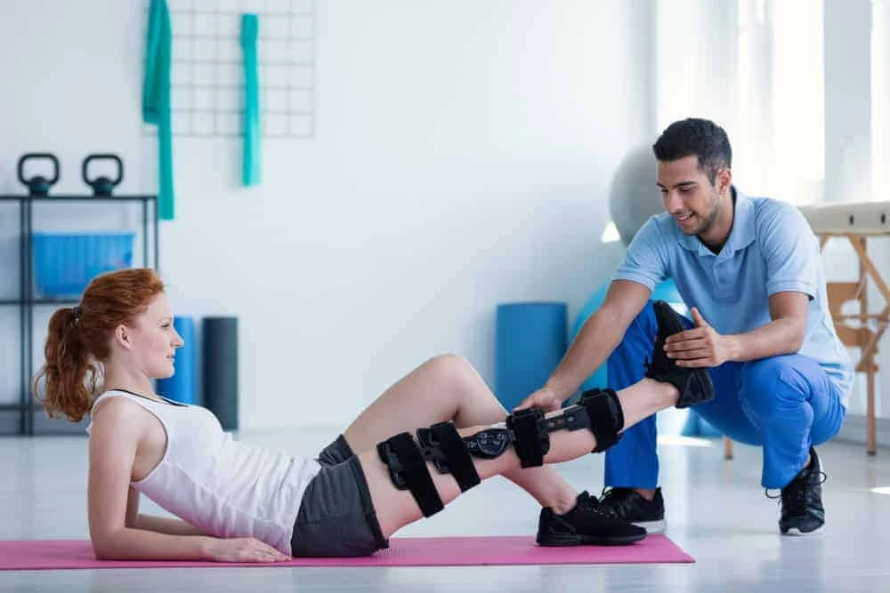 A woman with red hair lies on a pink yoga mat, wearing a knee brace. A man kneels beside her, assisting with a leg exercise in what looks like a physical therapy NYC session. The room, filled with kettlebells and a large ball, is bathed in bright natural lighting.