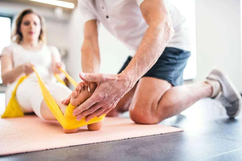 In a bright NYC room, a person kneels to assist another with an exercise, holding a yellow resistance band around their feet. The seated individual, dressed in white workout clothes on a fitness mat, embodies the essence of physical therapy NYC for effective pain management.
