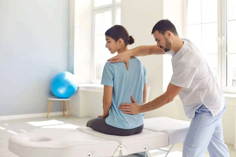 A physical therapist specializing in pain management NYC helps a woman sitting on a massage table by applying gentle pressure to her back. The room is bright with large windows, and an exercise ball is visible in the background, reflecting the holistic approach of physical therapy NYC.