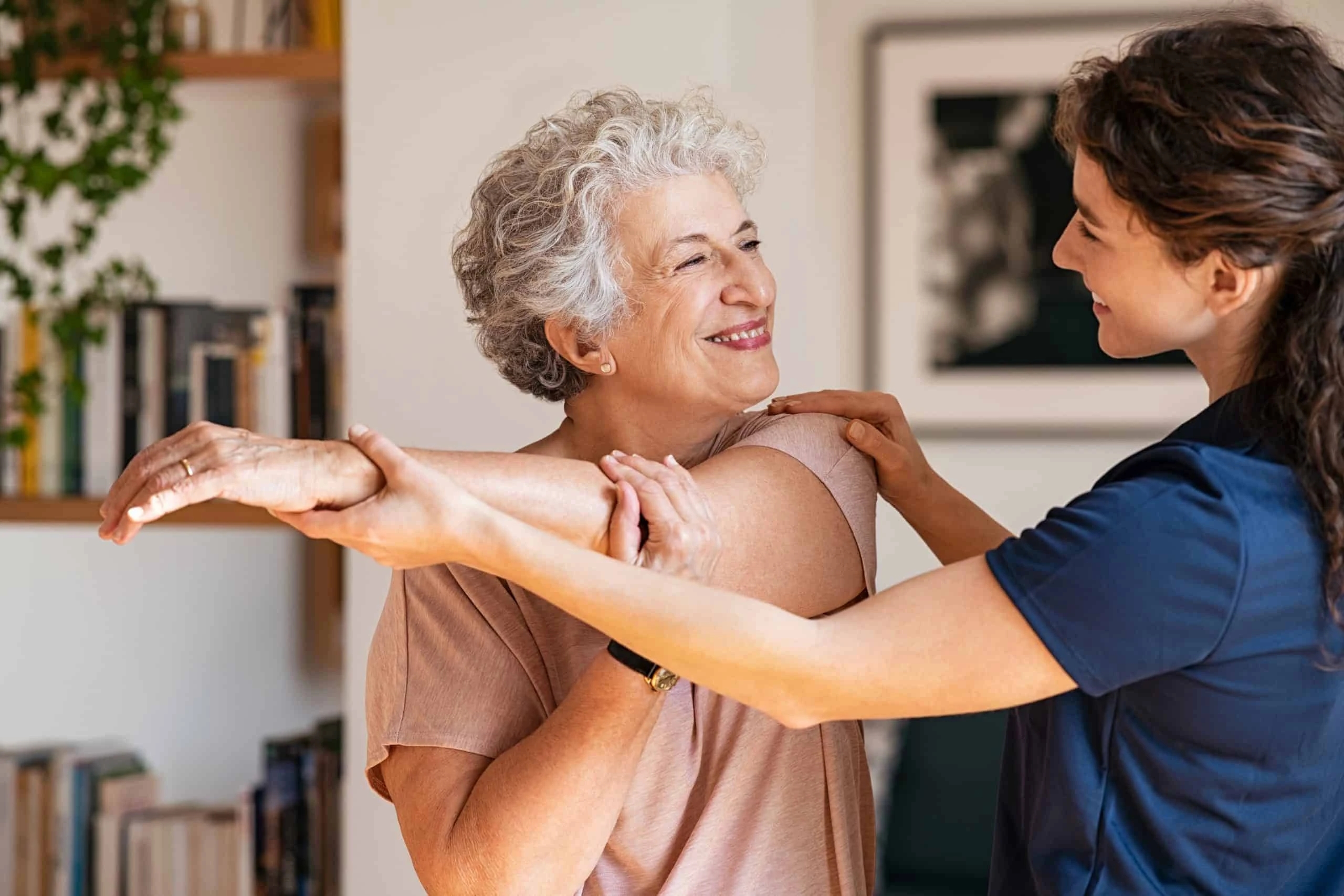 An elderly woman smiles as she stretches her arm with the assistance of a younger woman in a blue shirt, highlighting the benefits of physical therapy NYC. They are in a room with shelves and framed pictures in the background.