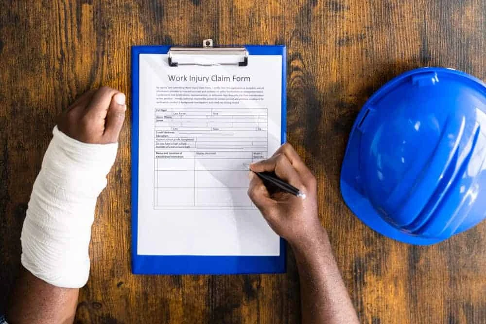 A person with a bandaged arm fills out a "Work Injury Claim Form" on a clipboard. A blue hard hat sits on the wooden table beside them, symbolic of their journey toward recovery and pain management in NYC.