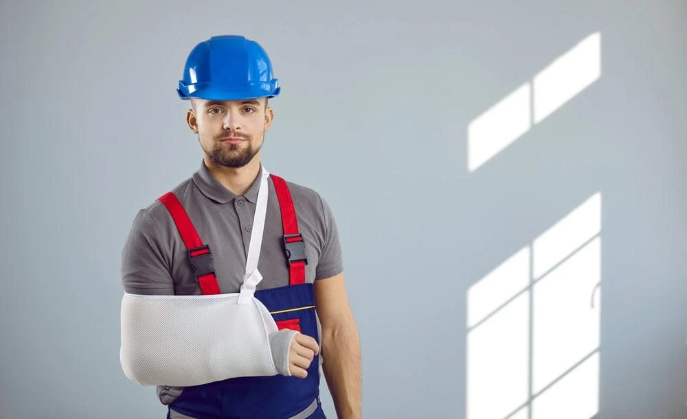 A construction worker, dressed in a blue hard hat and red suspenders, stands with his right arm in a white sling. He leans against a light-colored wall, shadows hinting at a nearby window. Recently starting physical therapy in NYC, he's optimistic about his recovery journey.