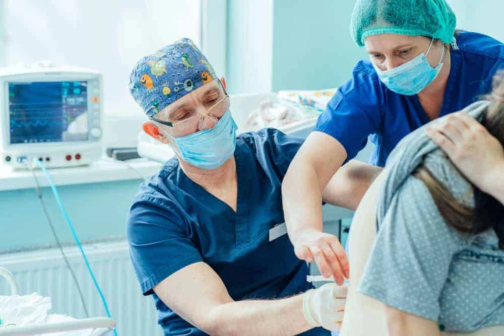 A healthcare professional in a cap and mask administers an injection to a patient, focusing on pain management. Another staff member assists, wearing a green hair cover. Medical equipment can be seen in the background of this NYC clinic.