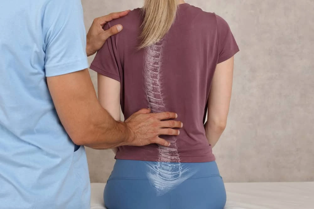 A person with long blonde hair sits with their back to the camera, wearing a maroon shirt featuring a spine image. Another's hand gently supports them, hinting at expert guidance in pain management NYC or physical therapy NYC.