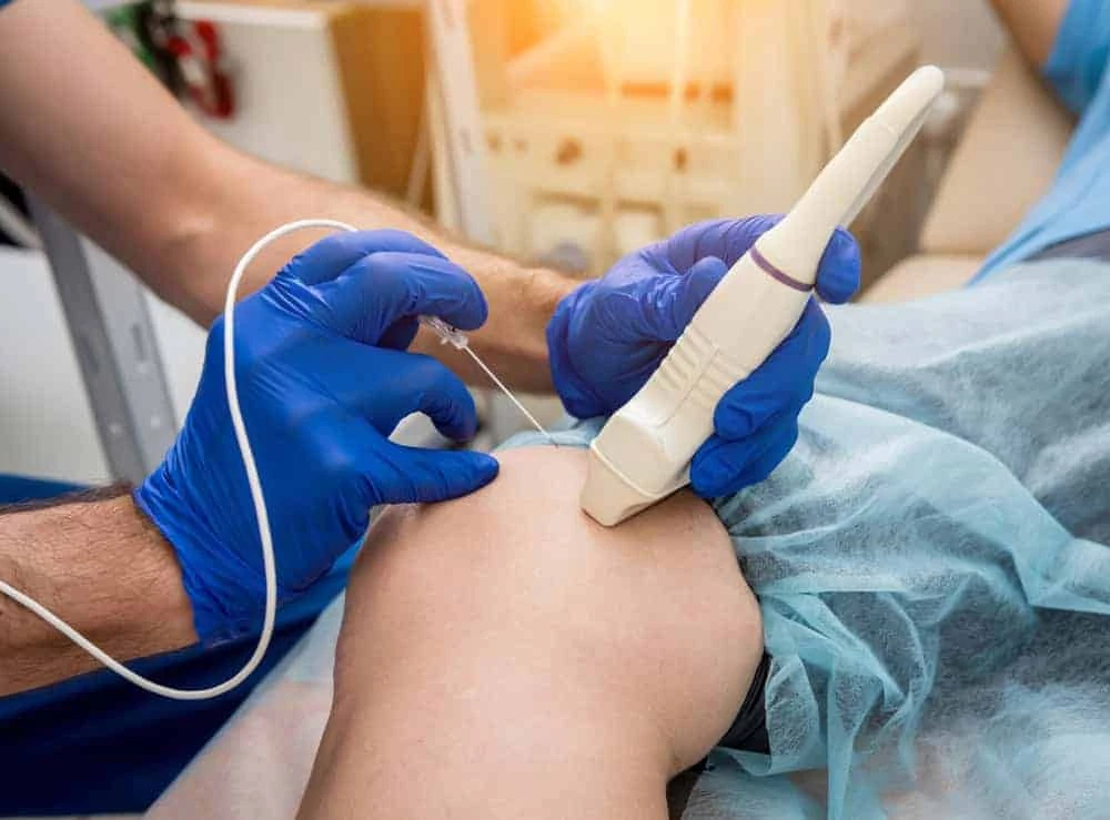 A medical professional in blue gloves conducts an ultrasound-guided knee injection for pain management NYC in a clinic. The patient, prepared with a sterile blue drape, receives expert care focused on alleviating discomfort and promoting recovery.