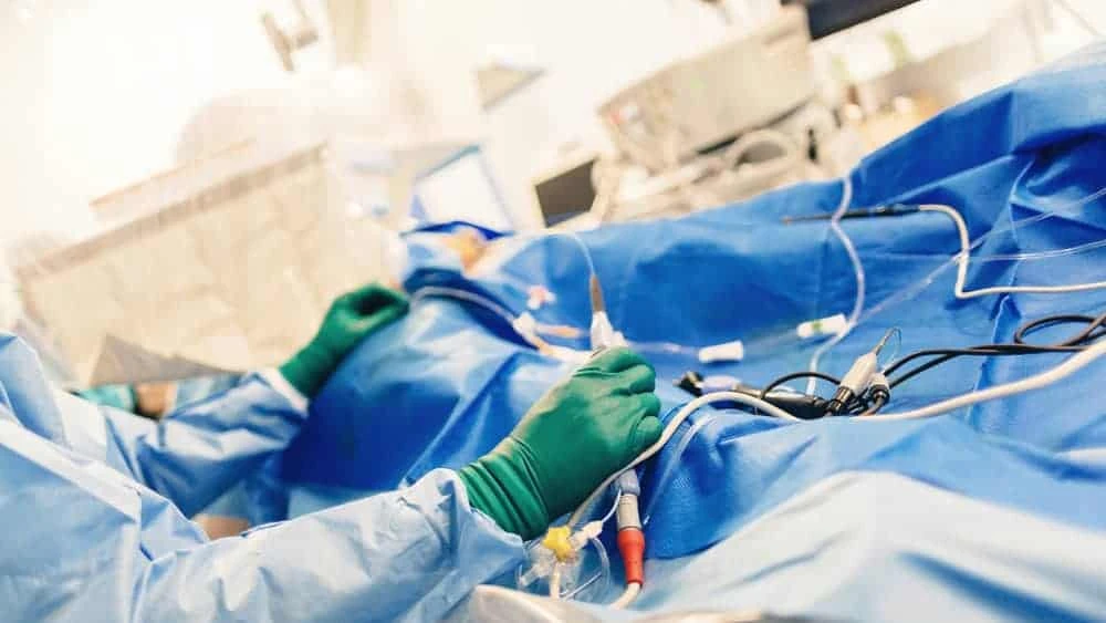 A surgeon in green gloves skillfully navigates between surgical tools while monitoring instruments during the procedure. The operating area is draped in blue, with medical equipment visible, seamlessly blending into the broader spectrum of pain management NYC practices.