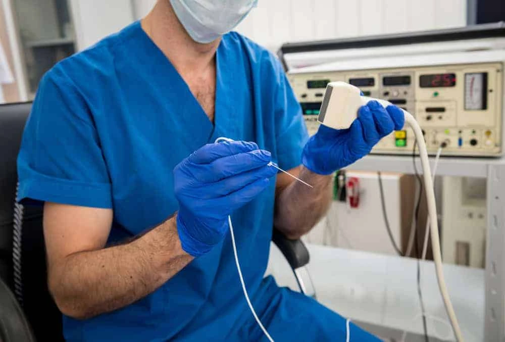 A medical professional in blue scrubs and gloves holds a small, square ultrasound probe and a thin needle. They are seated in a clinical setting with medical equipment and a monitor, specializing in pain management NYC for comprehensive care.
