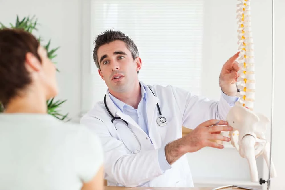 In a bright office, a doctor in a white coat and stethoscope explains spine anatomy to a patient using a spine model. The patient, interested in pain management NYC options, listens intently with their back to the camera.