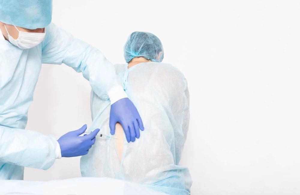 A healthcare professional in blue scrubs and gloves administers an injection into the back of a seated patient wearing similar attire, focusing on pain management NYC. Both are in a clinical setting with a plain white background.
