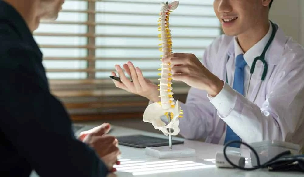 A doctor in a white coat and stethoscope sits at a desk, discussing pain management NYC options with a patient while explaining a model of the human spine. The room is bright with natural light streaming through the window blinds.