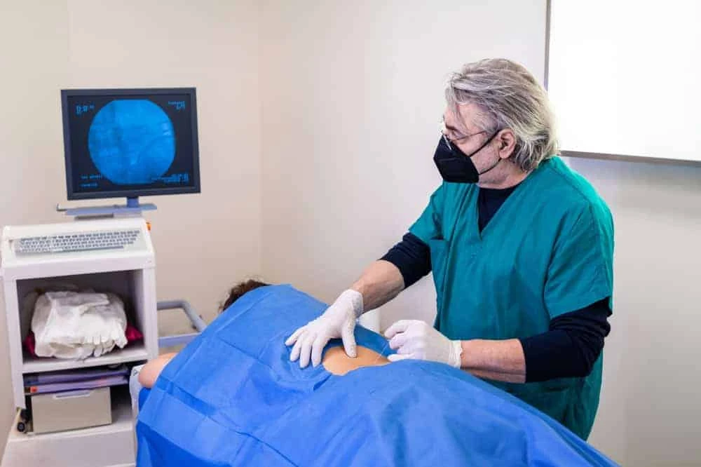 A healthcare professional in a green gown and black mask is performing a procedure on a patient lying face down, covered in blue sheets. A medical monitor displays diagnostic imaging in the background, all part of comprehensive physical therapy NYC offers for effective pain management.