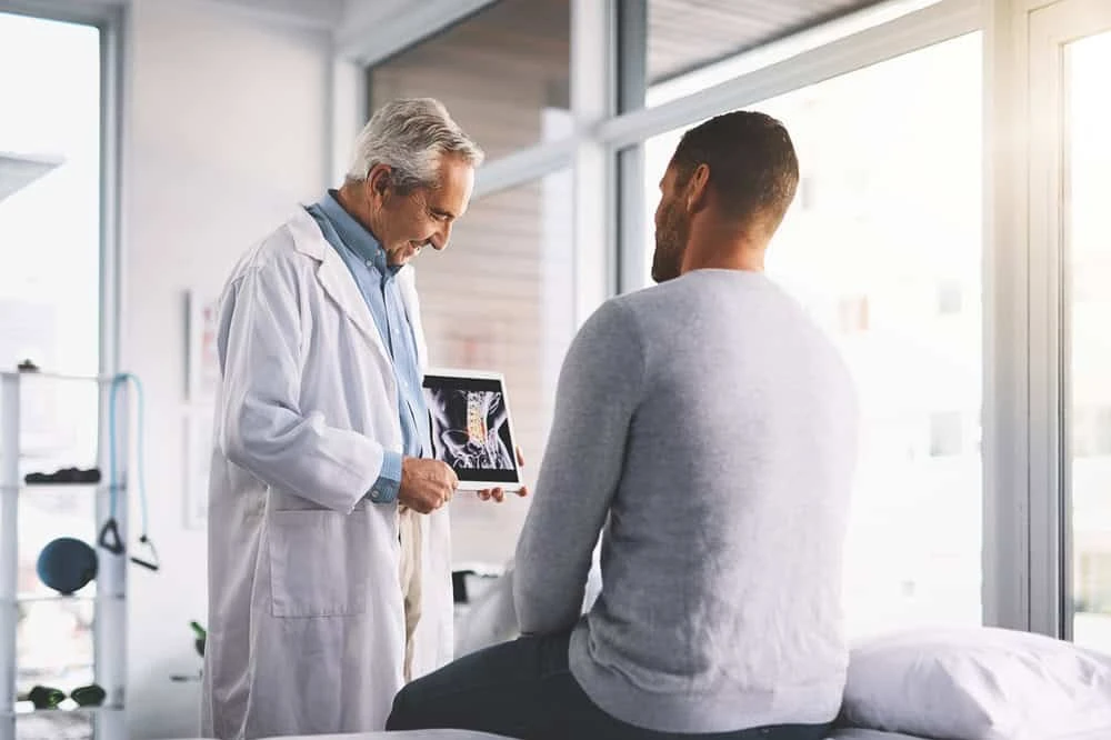A doctor in a white coat shows an X-ray to a patient sitting on a medical examination bed, discussing pain management options in NYC. The bright room with large windows enhances their engaged conversation, reflecting the dedication found in top physical therapy practices across the city.
