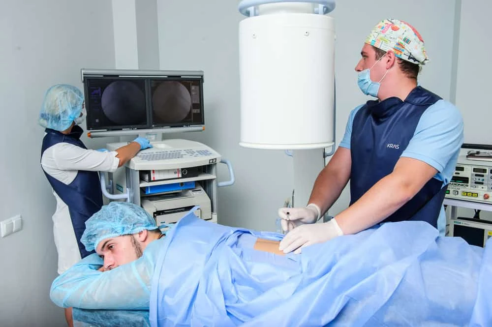 A medical professional in a surgical cap and mask performs a procedure focused on pain management NYC, as the patient lies face down on a table covered by a blue drape. Another specialist monitors screens displaying medical images, with equipment and monitors visible in the background.