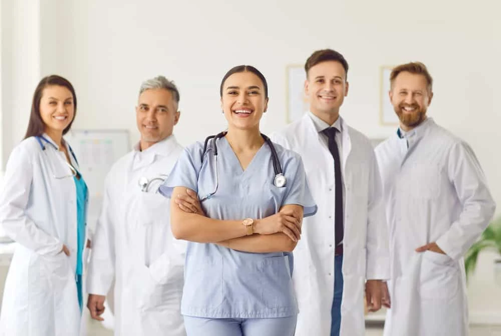 A group of five medical professionals stands together, smiling. The central figure is a woman in blue scrubs with her arms crossed. The others are in white lab coats, specializing in pain management NYC. The background is a bright, clinical setting.