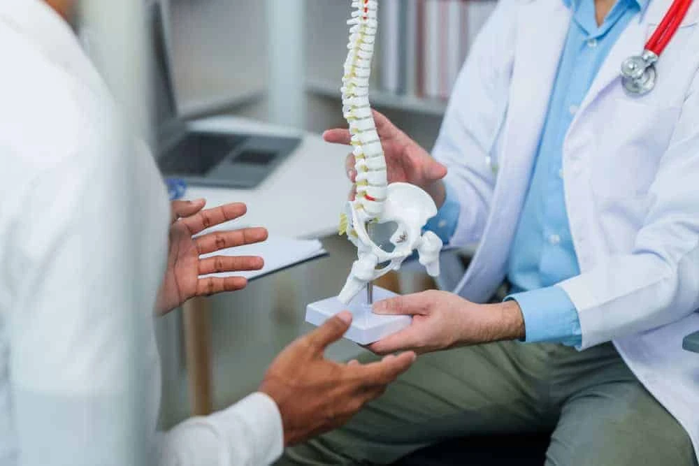 In a medical office, a doctor in a white coat holds a model of a human spine, explaining the intricacies of pain management NYC-style to another person. A laptop rests on the desk, hinting at advanced physical therapy NYC options available for effective treatment plans.