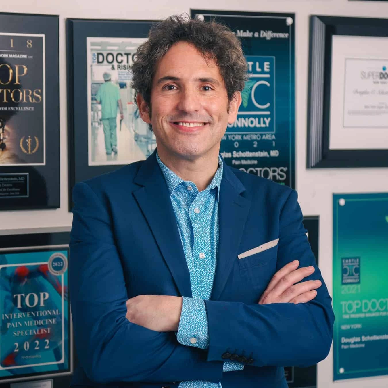 A man in a blue suit with arms crossed smiles in front of a wall displaying framed awards and certificates, highlighting his expertise in physical therapy NYC.