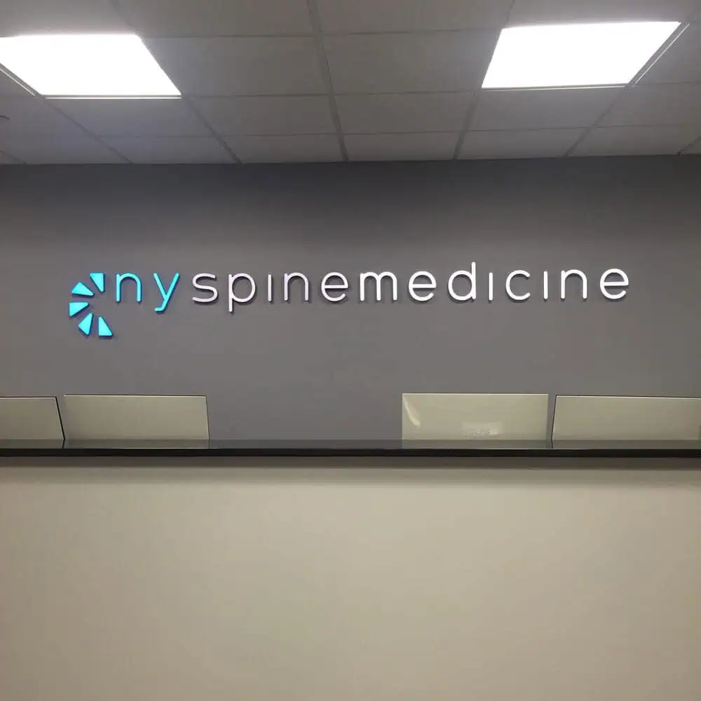 Wall signage displaying the text "nyspinemedicine" in lowercase, with "ny" in light blue and "spinemedicine" in white. A circular, segmented logo in light blue sits to the left, reflecting their expertise in pain management NYC.