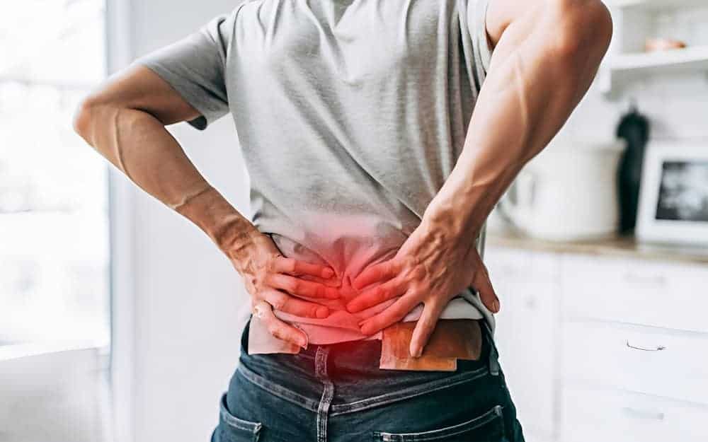 A person wearing a gray shirt and jeans stands in a kitchen, holding their lower back with both hands. A red area highlights discomfort, suggesting they might benefit from pain management NYC solutions.