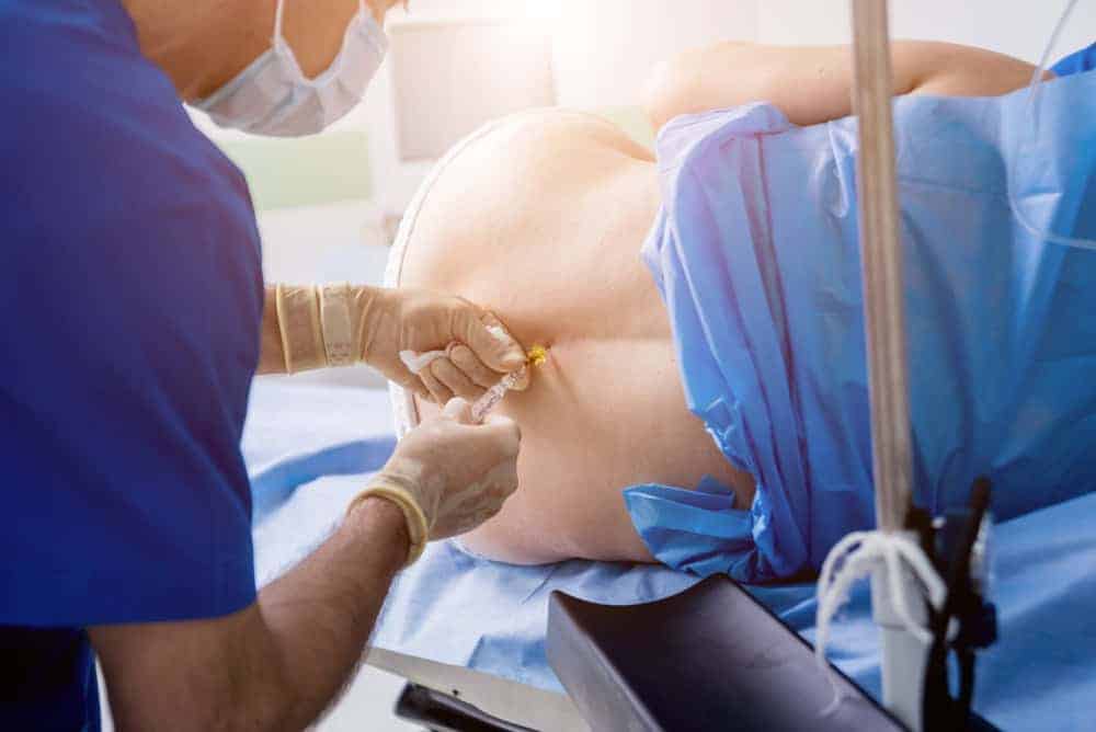 A medical professional in blue scrubs administers an epidural to a patient lying on their side, covered with a blue drape. The healthcare worker wears gloves and a mask in this pain management NYC setting, emphasizing precision and care in the clinical environment.