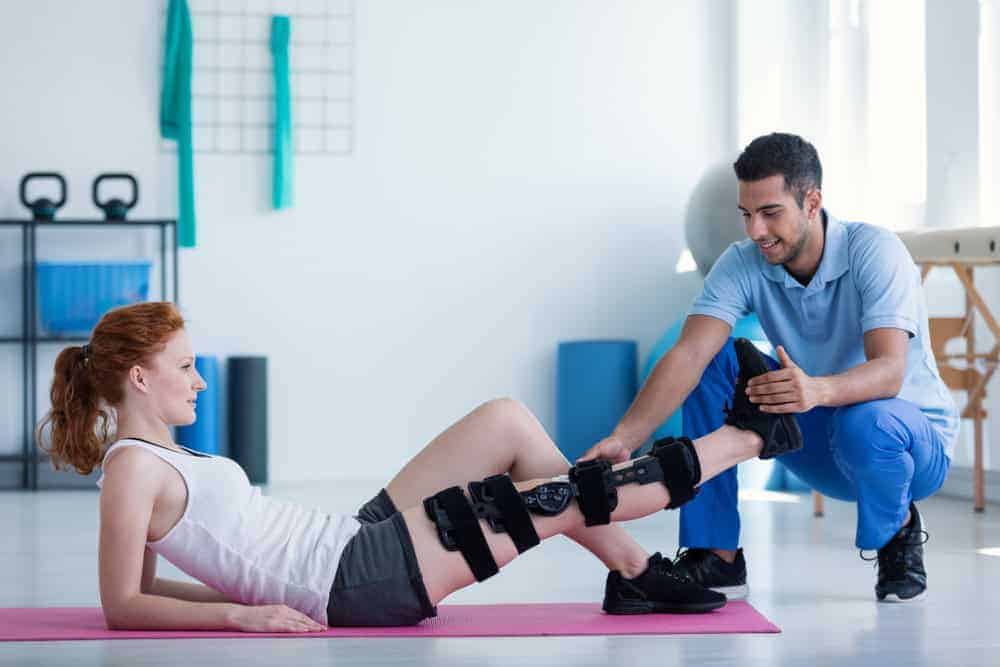 A woman with red hair lies on a pink yoga mat, wearing a knee brace. A man kneels beside her, assisting with a leg exercise in what looks like a physical therapy NYC session. The room, filled with kettlebells and a large ball, is bathed in bright natural lighting.