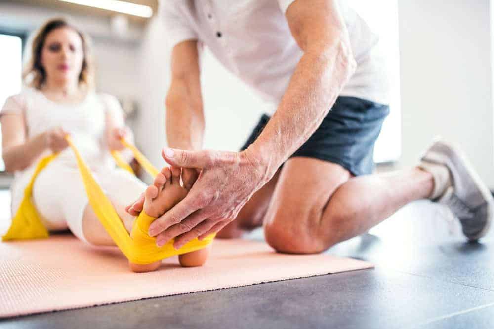 In a bright NYC room, a person kneels to assist another with an exercise, holding a yellow resistance band around their feet. The seated individual, dressed in white workout clothes on a fitness mat, embodies the essence of physical therapy NYC for effective pain management.