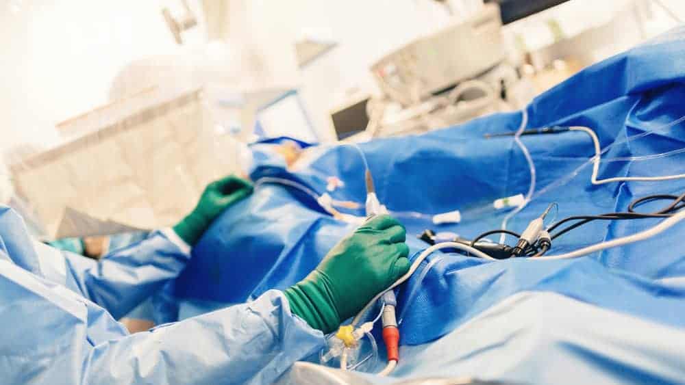 A surgeon in green gloves skillfully navigates between surgical tools while monitoring instruments during the procedure. The operating area is draped in blue, with medical equipment visible, seamlessly blending into the broader spectrum of pain management NYC practices.