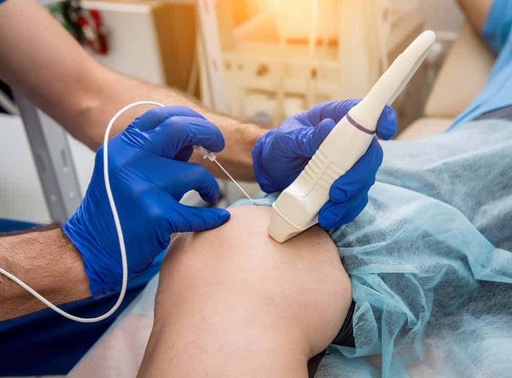 A medical professional in blue gloves conducts an ultrasound-guided knee injection for pain management NYC in a clinic. The patient, prepared with a sterile blue drape, receives expert care focused on alleviating discomfort and promoting recovery.
