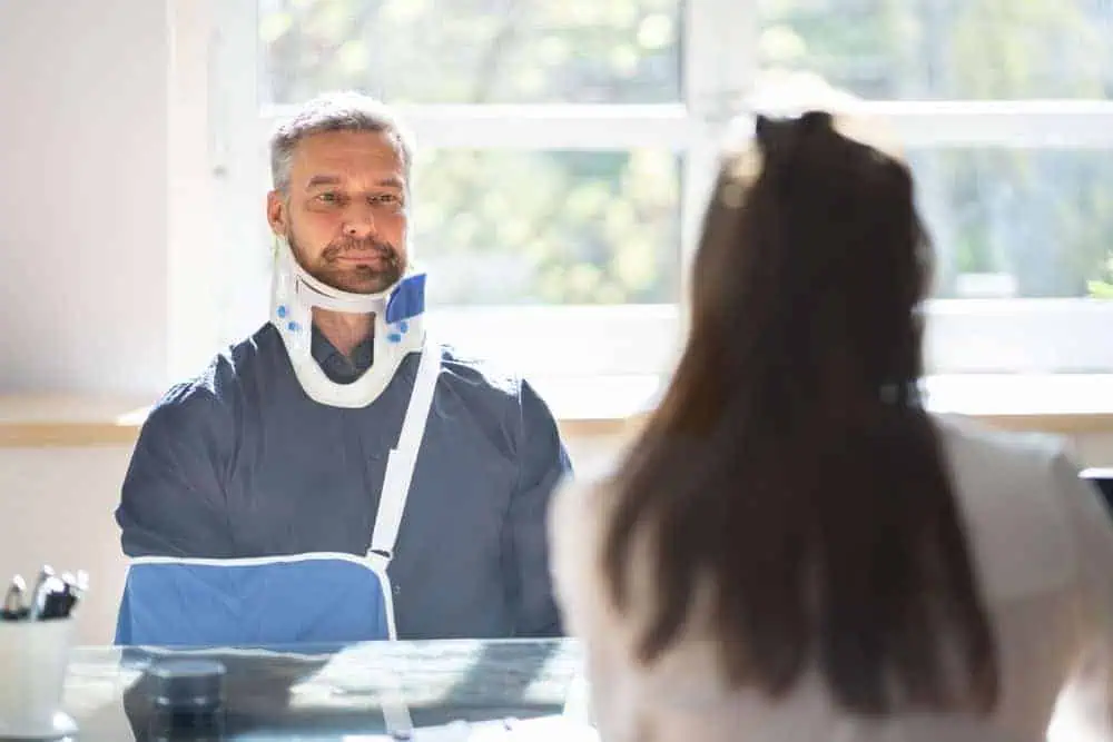 A man with a neck brace and arm sling sits across from a woman at a desk, likely discussing pain management in NYC. Sunlight streams through the window behind them. The man looks serious, while the woman's back faces the camera. Papers and pens are scattered on the desk.