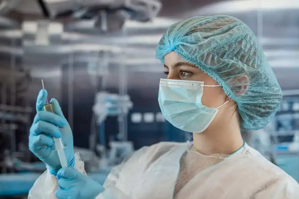 A healthcare worker in a blue surgical cap, mask, and gloves is holding a syringe with a focused expression, embodying the precision vital for pain management NYC. The background is a blurred medical setting.