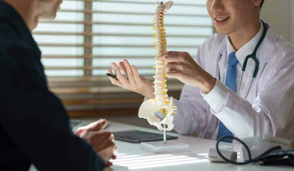 A doctor in a white coat and stethoscope sits at a desk, discussing pain management NYC options with a patient while explaining a model of the human spine. The room is bright with natural light streaming through the window blinds.