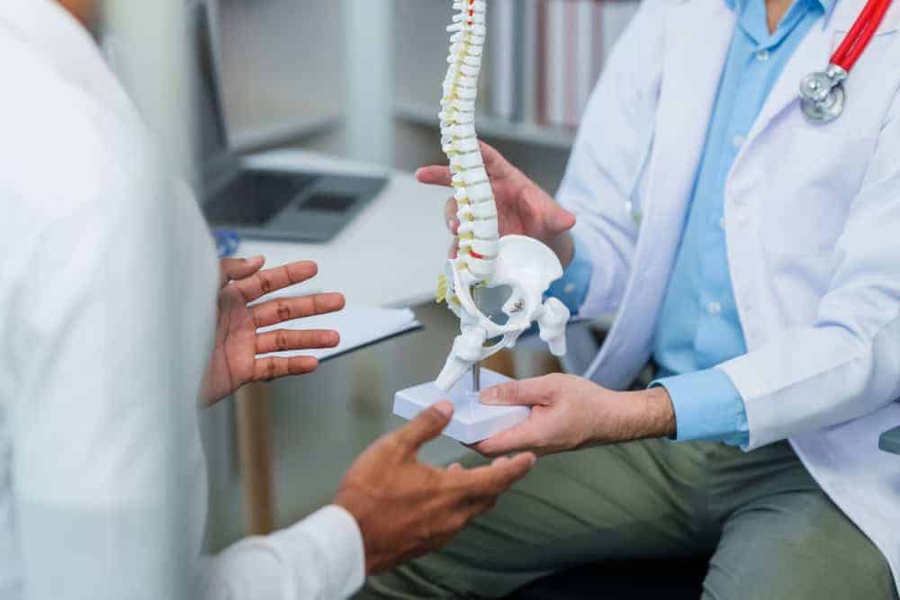 In a medical office, a doctor in a white coat holds a model of a human spine, explaining the intricacies of pain management NYC-style to another person. A laptop rests on the desk, hinting at advanced physical therapy NYC options available for effective treatment plans.