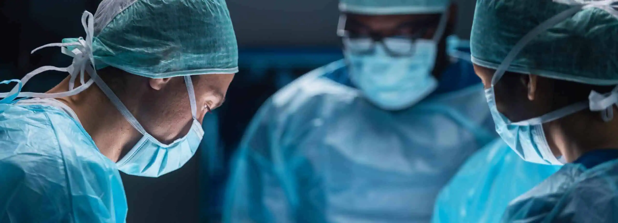 Three surgeons are focused on a procedure in an operating room, embodying the dedication that complements holistic care like physical therapy NYC. Draped in blue surgical gowns, masks, and hair caps, they're under bright surgical lights in this concentrated and sterile environment.