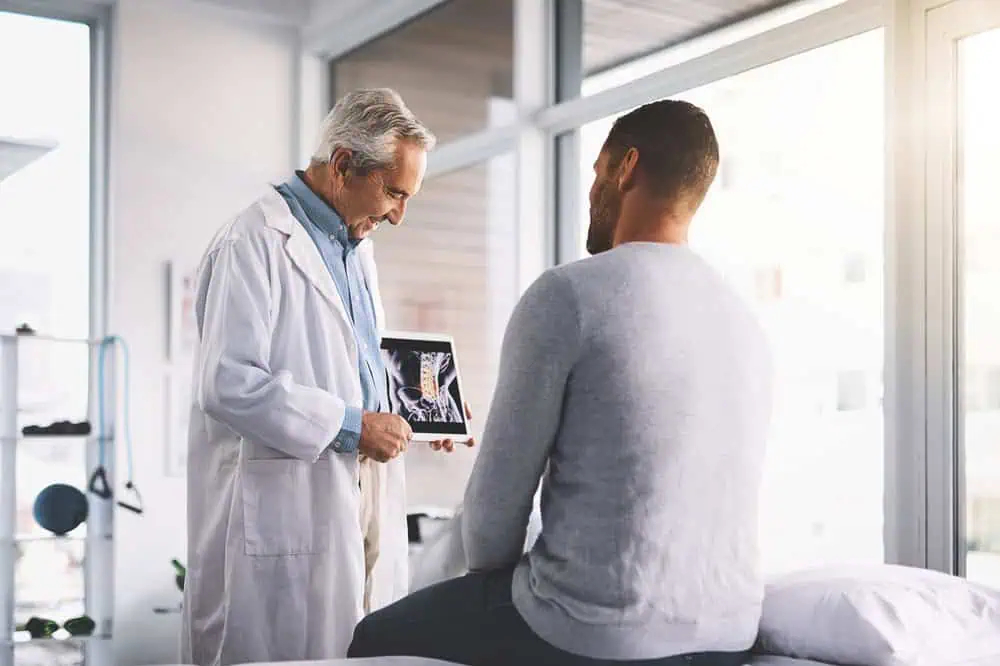 A doctor in a white coat shows an X-ray to a patient sitting on a medical examination bed, discussing pain management options in NYC. The bright room with large windows enhances their engaged conversation, reflecting the dedication found in top physical therapy practices across the city.