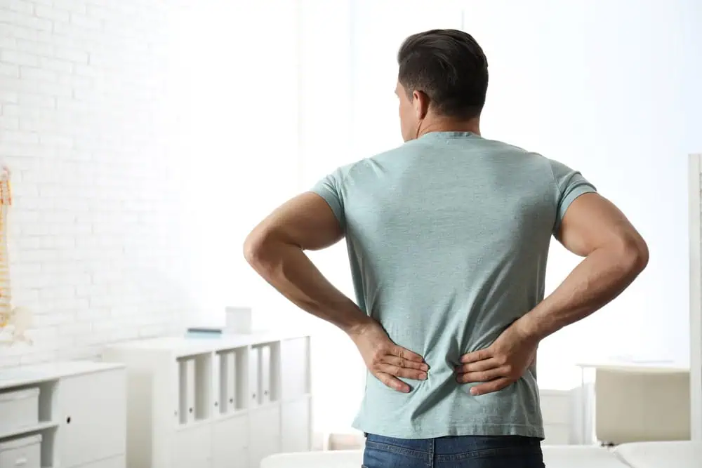 A man in a light gray t-shirt and jeans stands with his back to the camera, holding his lower back, perhaps contemplating physical therapy NYC as he seeks relief in a bright room with white walls and shelves.