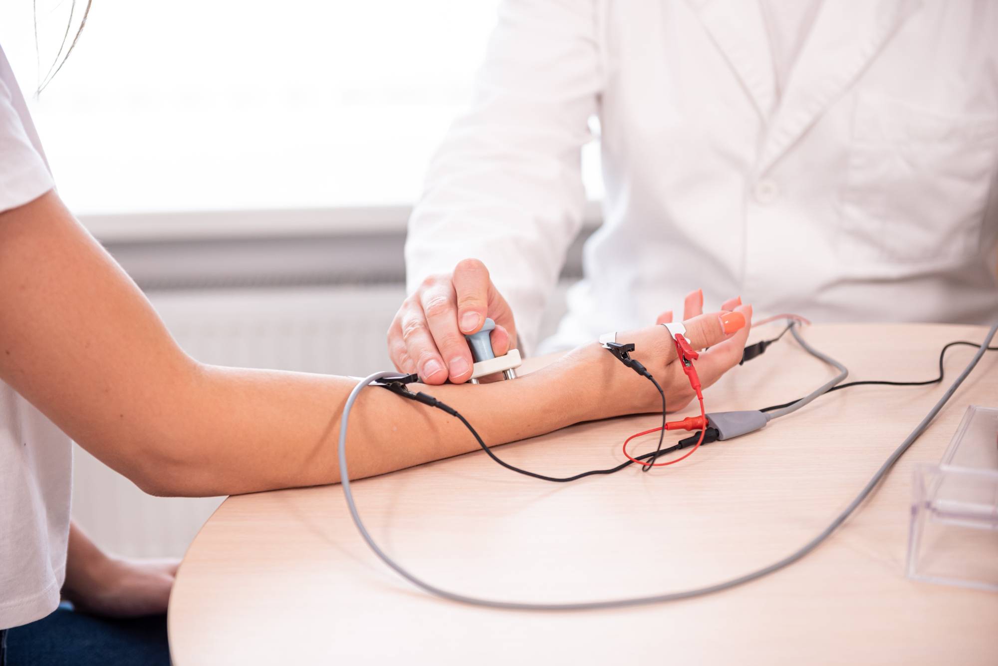 A doctor in a white coat is conducting a medical test on a patient’s arm using sensors and wires attached to the patient's hand and wrist, likely performing a nerve or muscle function test.