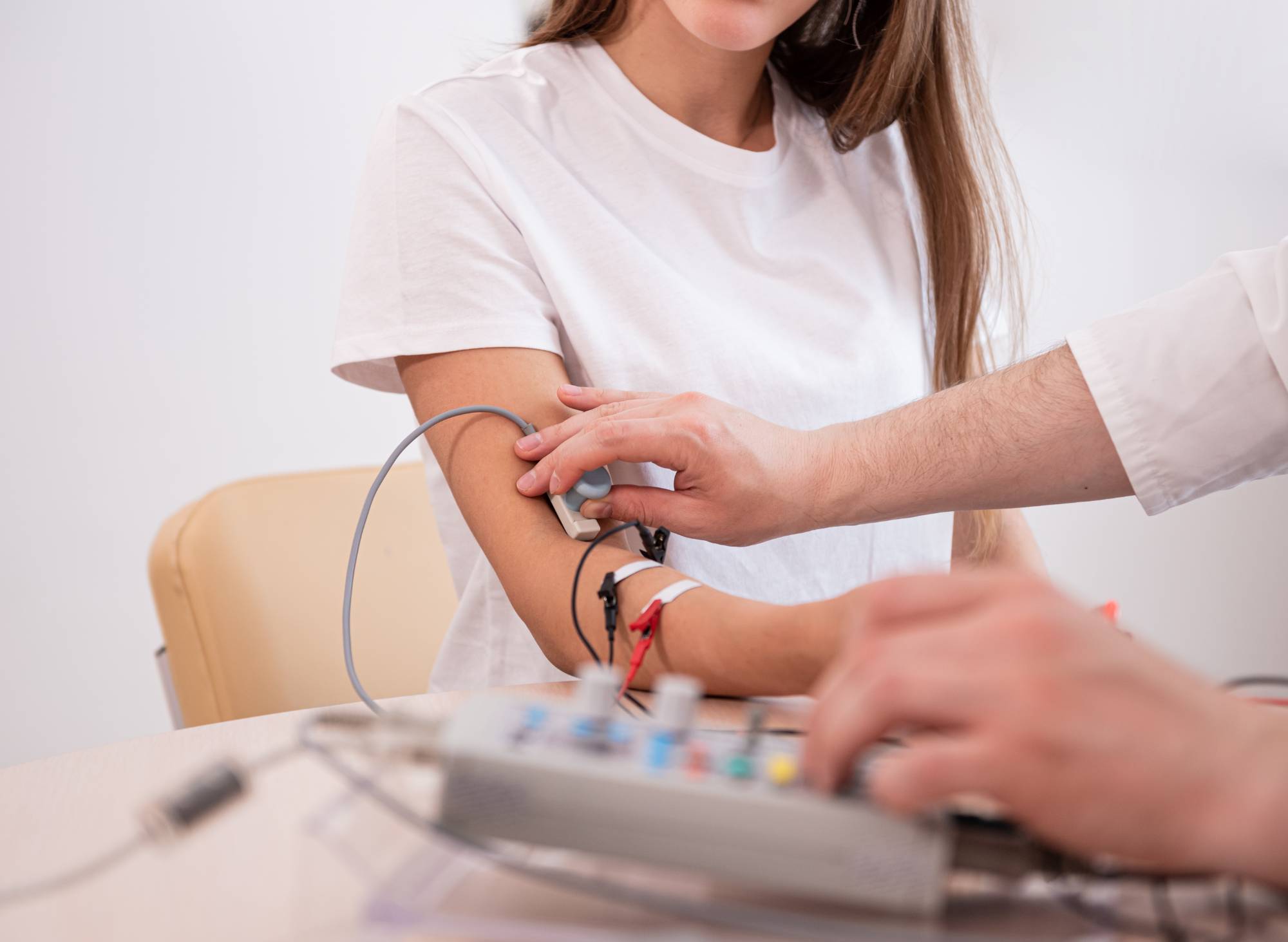 A person wearing a white t-shirt is seated while another person attaches sensors to their arm, which are connected to a polygraph machine on the table.