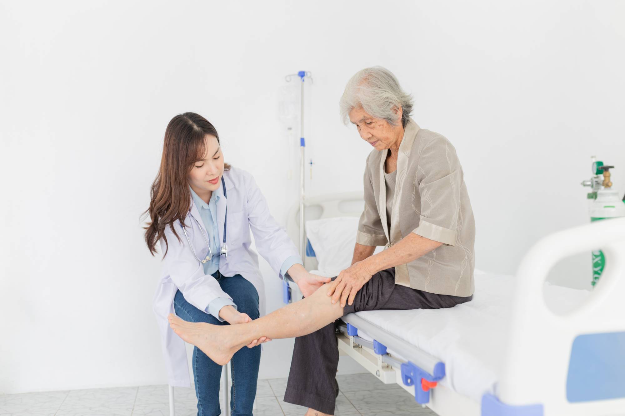 A doctor examines the lower leg of an elderly woman sitting on a hospital bed. The doctor gently holds the patient’s leg, while the patient looks down at her leg. Medical equipment is visible in the background.