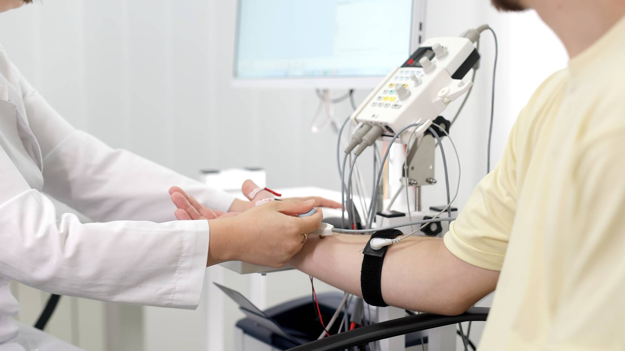 A healthcare professional attaches electrodes and medical devices to a patient's arm, likely for a diagnostic test or monitoring, in a clinical setting with medical equipment visible in the background.